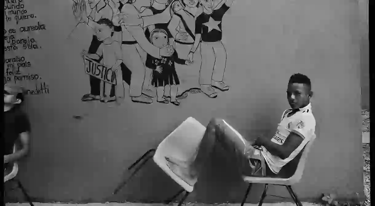 Tenosique, Mexico. A resident of La 72 shelter waits for mealtime in front of one of the many murals painted by volunteers and residents. The painting of a group of marchers demanding justice and freedom sits alongside a poem by Mario Bendetti, a Uruguayan writer forced to flee his country after a military coup in 1973.