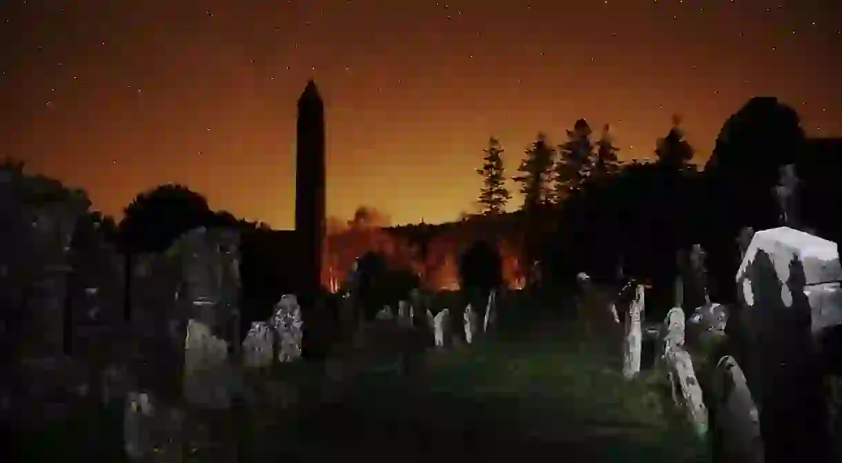 The round tower and graveyard at Glendalough at night, with the glow of Dublin to the north