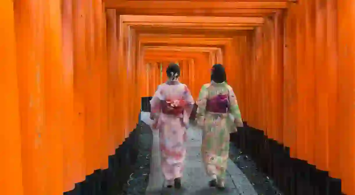 Two geishas among red wooden Tori Gate at Fushimi Inari Shrine in Kyoto, Japan.