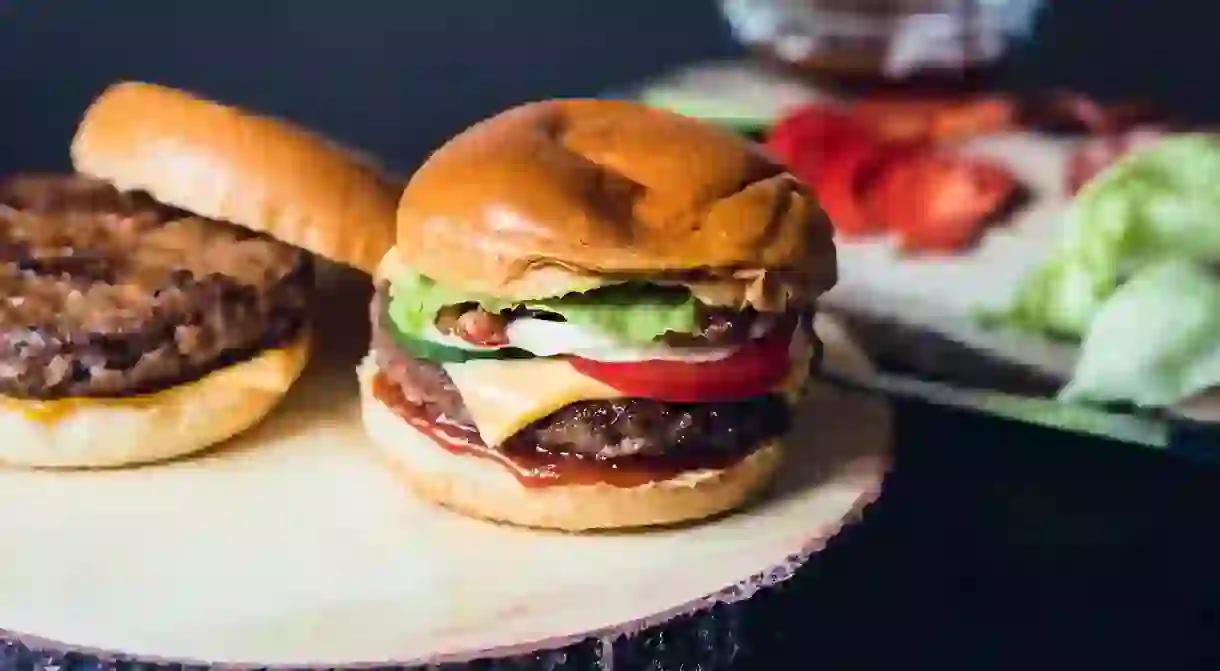 Self made cheeseburgers on a wooden plate with ingredients in the background