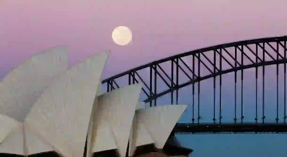 M24KCA A full moon hovers over the Sydney Harbour Bridge and Opera House. The sky glows pink and blue as the sunrises.