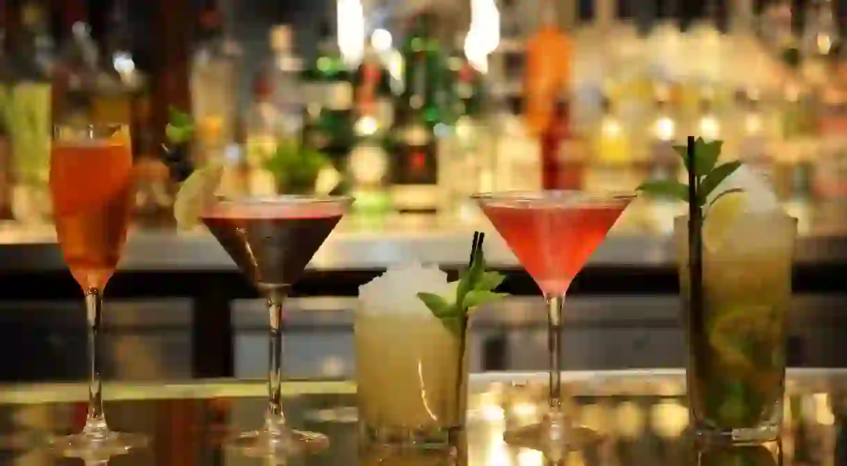 A row of colourful cocktails on a bar, with other drinks behind.