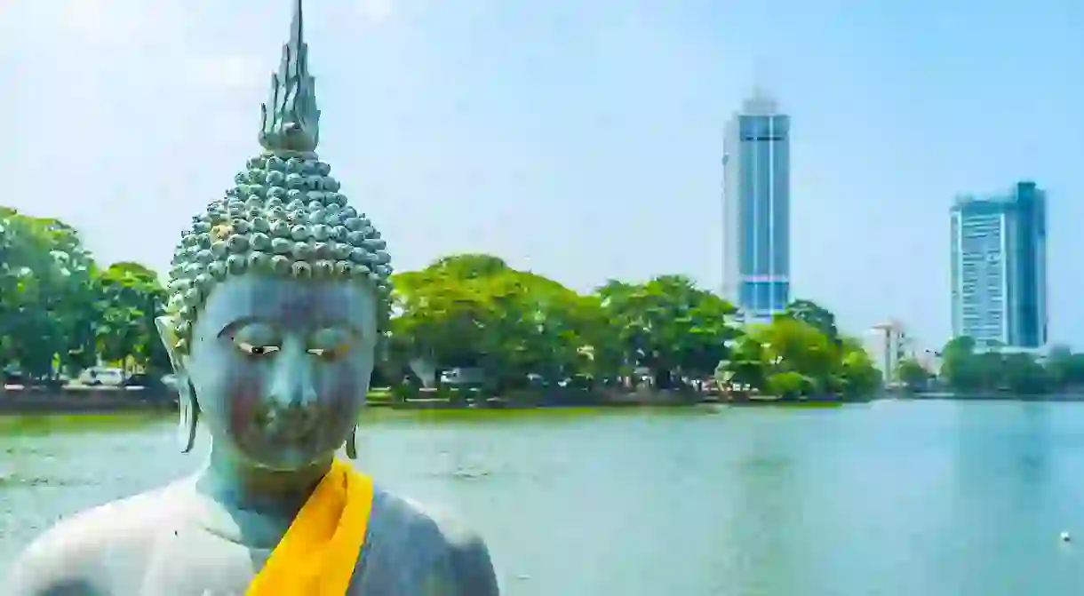 The bronze Buddha statue in Seema Malaka Temple with the surface of Beira lake and modern skyscrapers on background, Colombo, Sri Lanka.