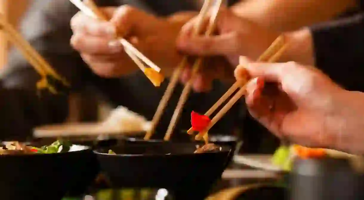 Four sets of chopsticks grabbing Asian food out of various black bowls sitting on a table in a Thai food restaurant. Three pairs of chopsticks have food between them, and one is in a black bowl. There is a persons chest out of focus in the background. There are four sets of chopsticks visible, but there are only three hands visible. There is a set of chopsticks coming into view from the left. The hand in front is in focus, while the two hands in the background are slightly blurred.