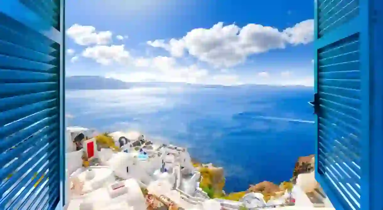 Hillside view through an open window with blue shutters of the caldera, sea and white village of Oia on the island of Santorini, Greece.