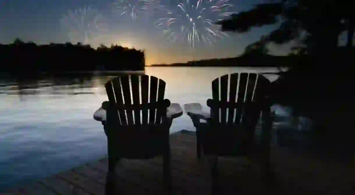 Canada Day celebration fireworks light up the sky over Muskoka Lake, Ontario, as two Adirondack chairs on the dock offer a front-row seat to the spectacular show.