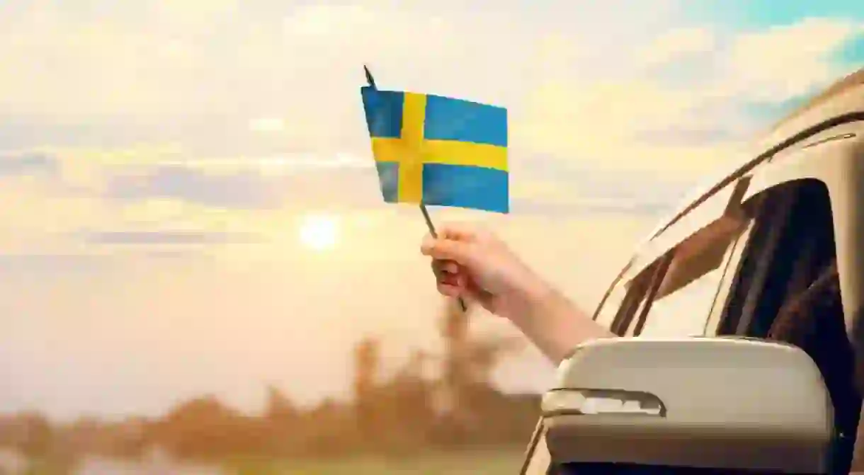 Waving the Sweden flag against the sunrise or sunset from a car driving along a country road. Holding the Sweden flag, traveling by car, on a weekend trip.