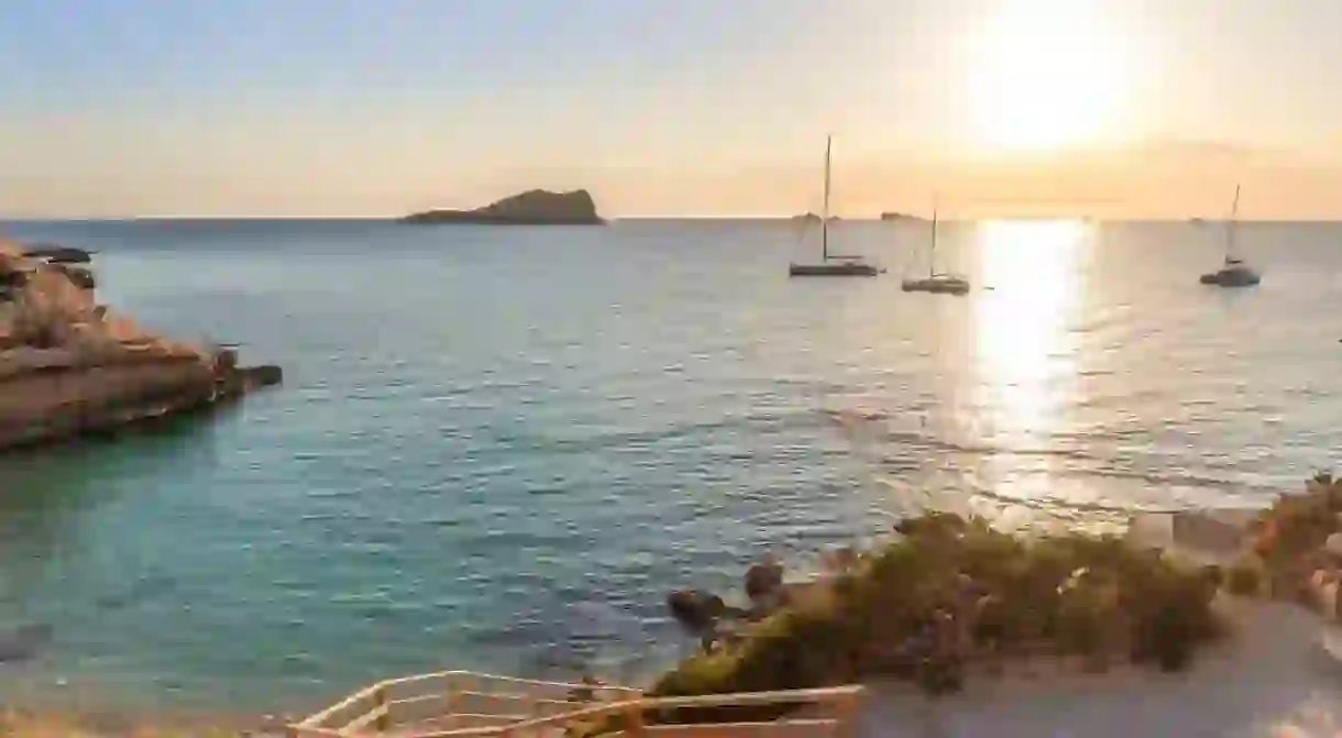 Sunset at Cala Conta beach in Ibiza, Spain, with emerald clear water and sailboats on the horizon.