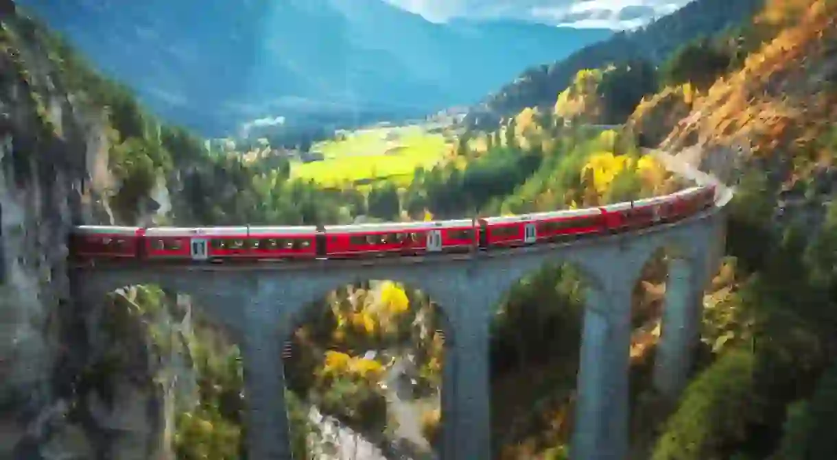 Aerial view of modern red train on Landwasser viaduct in alpine mountains, orange forest at sunset in autumn. Bernina Express, Switzerland.
