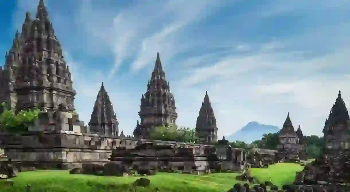 Ancient stone ruins on green field and Candi Prambanan or Rara Jonggrang, Hindu temple compound on background. Impressive architectural site. Yogyakarta, Central Java, Indonesia. Panoramic view.
